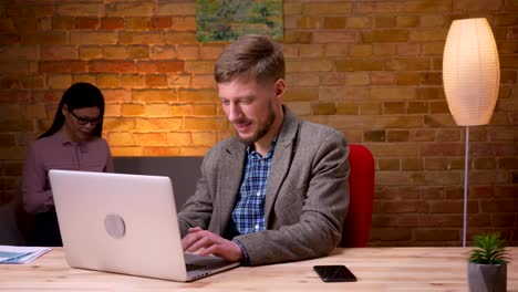 Closeup-shoot-of-adult-businessman-using-the-laptop-receiving-a-notification-on-the-phone-smiling-and-sharing-good-news-with-colleagues.Female-employee-using-the-cellphone-on-the-background