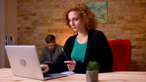 Closeup-shoot-of-adult-caucasian-businesswoman-working-on-the-laptop-indoors-in-the-office.-Businessman-handling-her-papers-sitting-the-couch-and-using-the-tablet-on-the-background