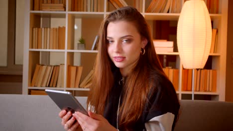 Portrait-of-young-teenage-girl-working-with-tablet-sitting-on-sofa-on-bookshelves-background-watches-into-camera-at-home.