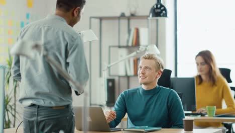 Handsome-Caucasian-IT-Specialist-Sitting-at-His-Desk-works-on-a-Laptop,-Has-Discussion-with-Indian-Project-Manager.-Diverse-Team-of-Young-Professionals-Working-in-Office