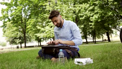 Un-joven-empresario-sonriente-sentado-en-el-parque-durante-el-almuerzo-y-escribiendo-en-la-tableta