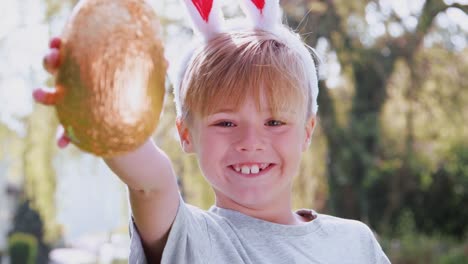 Portrait-of-boy-wearing-bunny-ears-on-Easter-egg-hunt-outdoors-at-home-holding-up-chocolate-egg-to-camera-and-smiling---shot-in-slow-motion