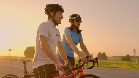 Ein-Mann-und-eine-Frau-in-Helmen-mit-Fahrrädern-stehen-und-reden-bei-Sonnenuntergang.-Ruhe-nach-einer-Radtour-auf-der-Autobahn.-Rennräder.-Paar-Sport.