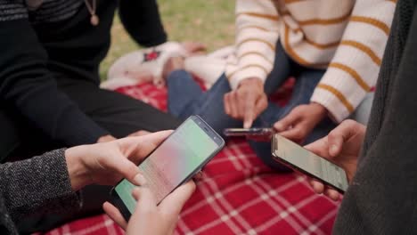 Close-up-of-friends-sitting-together-on-red-blanket-using-mobile-app-in-the-park