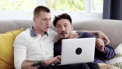 Gay-couple-relaxing-on-couch-using-laptop-computer.-Thinking-and-typing.