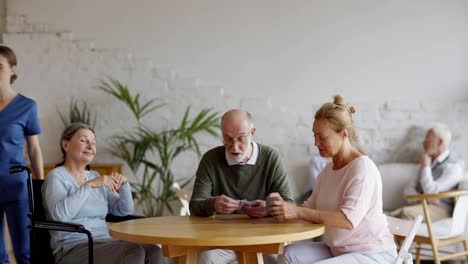 Betreuerschieben-Rollstuhl-von-behinderten-alten-Frau-zu-helfen,-sie-mit-älteren-Freunden-Spielen-Karten-am-Tisch-im-Pflegeheim.-Älterer-Mann-und-Frau-sprechen-auf-Sofa-im-Hintergrund