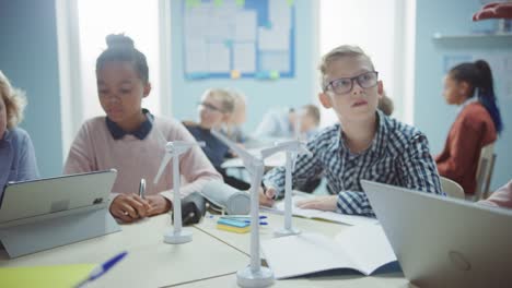 In-der-Grundschule:-Klasse-der-brillanten-kleinen-Kinder-arbeiten-als-Team-mit-Tablet-Computern,-um-Windkraftanlagen-zu-programmieren.-Klassenzimmer-mit-Kindern-Lernen-über-umweltfreundliche-Formen-erneuerbarer-Energie