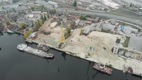 Aerial-view-of-industrial-part-of-the-city-with-cargo-cranes-and-a-pile-of-sand-with-barge-parked-on-the-bank-of-the-river.-Industrial-concept