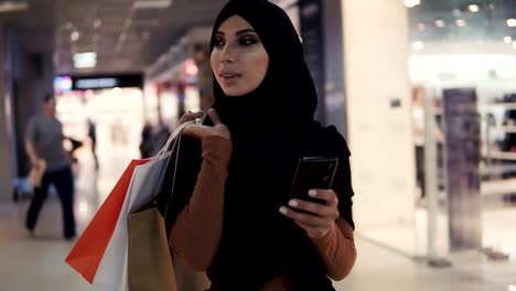 Attraktive-muslimische-Frau-trägt-schwarze-Hijab-Kopftuch-zu-Fuß-in-der-Shopping-Mall-und-mit-dem-Smartphone.-Kommunikation,-Online-Shopping,-Social-Network-Konzept