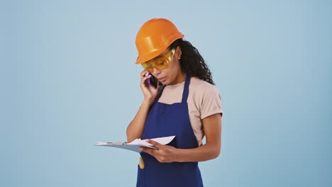 Dark-skinned-woman-industrial-worker-or-builder-in-hard-hat-and-protective-goggles-is-talking-by-smartphone-and-looking-at-papers-on-clipboard