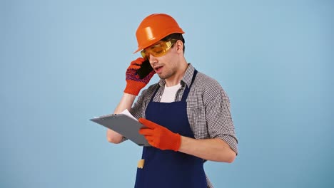 Young-guy-in-safety-wear,-hard-hat,-protective-goggles,-gloves.-Talking-by-mobile-phone,-looking-at-papers-on-clipboard.-Posing-on-blue-background