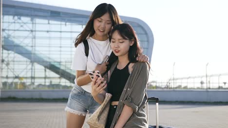 Hermosas-chicas-asiáticas-de-25-años-con-el-pelo-largo-mirando-en-el-teléfono-y-sonriendo-cerca-del-moderno-edificio-del-aeropuerto