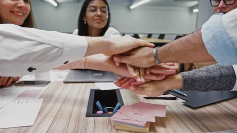 Team-of-diverse-colleagues-putting-their-hands-on-top-of-each-other-after-work-on-business-project,-sitting-at-table-in-office