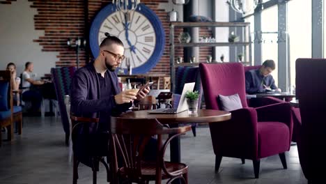 A-young-hipster-drinks-a-cocktail-while-sitting-at-a-wooden-table-in-a-restaurant,-a-man-looks-through-incoming-calls-on-a-mobile-phone,-in-front-of-him-is-a-laptop-for-work