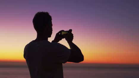 Mann,-der-Panorama-Foto-mit-Handy-am-Strand-bei-Sonnenuntergang