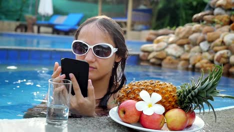 Mujer-hermosa-joven-utiliza-teléfono-móvil-en-la-piscina-al-lado-del-cristal-de-la-placa-con-frutas-tropicales-en-cámara-lenta-y-el-agua.-HD,-1920-x-1080