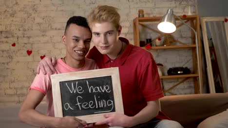 A-happy-international-gay-couple-is-sitting-on-the-couch-and-holding-a-sign.-We-have-feeling.-Look-at-the-camera.-Home-comfort-on-the-background.-60-fps