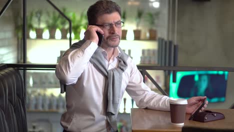 Mature-man-in-a-white-shirt-with-sweater-in-eyeglasses-looks-at-the-tablet-and-speaks-on-the-phone-comparing-data