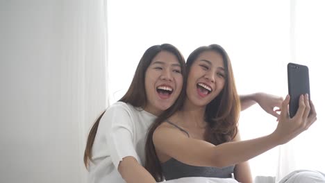 Hermosas-mujeres-asiáticas-jóvenes-LGBT-lesbiana-feliz-pareja-sentada-en-el-abrazo-de-la-cama-y-usar-teléfono-tomando-selfie-dormitorio-juntos-en-casa.-Lesbianas-LGBT-pareja-juntos-en-el-interior-concepto.-Pasar-tiempo-agradable-hogar.