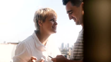 Happy-young-multiethnic-romantic-couple-standing-at-a-small-sunny-balcony-using-smartphone-social-network-apps-smiling