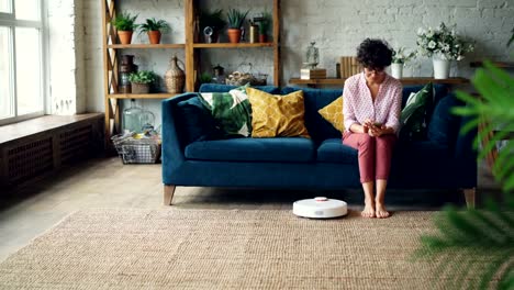 Attractive-woman-is-turning-on-robotic-vacuum-cleaner-sitting-on-sofa-then-using-smartphone-and-resting-while-device-is-cleaning-the-house.-Technology-and-apartments-concept.