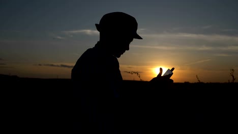 Silhouette-eines-jungen-verwendet-eine-Tablette-bei-Sonnenuntergang-im-Feld-liest-etwas-auf-dem-tablet