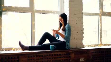 Fröhliche-junge-Frau-nutzt-Smartphone-sitzt-auf-der-Fensterbank-mit-Tasse-Tee-zu-Hause-entspannen-und-genießen-modernen-Technologie.-Menschen-und-Geräte-Konzept.