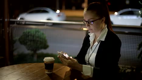 Schöne-junge-kaukasische-Frau-sitzt-nach-der-Arbeit-am-Abend-in-einem-Straßencafé-beim-Kaffee-und-plaudert-in-sozialen-Netzwerken-mit-einem-Smartphone.