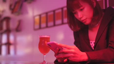 Young-asian-millennial-woman-drinking-beer-and-using-smartphone-in-nightclub-bar-at-night,-neon-pink-background