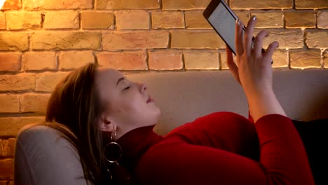 Closeup-shoot-of-young-pretty-plump-caucasian-female-typing-on-the-tablet-while-lying-on-the-couch-then-looking-at-camera-and-smiling