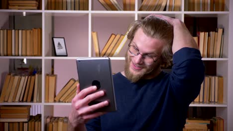 Closeup-shoot-of-adult-attractive-male-student-having-a-video-call-on-the-tablet-in-the-university-library-indoors