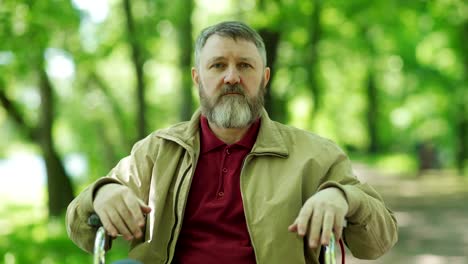 Medium-shot-of-disabled-mature-man-with-beard-sitting-in-wheelchair-and-looking-at-camera-in-green-park-on-windy-day