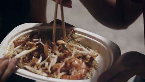 Asian-young-women-friends-travel-in-Bangkok,-Thailand,-beautiful-female-feeling-happy-walking-and-eating-Pad-Thai-at-The-Khao-San-Road.-Women-travel-eat-street-food-concept.-Slow-motion-shot.