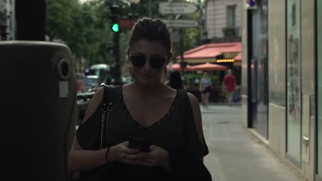 Atractiva-mujer-caucásica-con-gafas-de-sol,-pecas,-piercings-y-pelo-rojo-escribiendo-un-mensaje-de-texto-en-su-smartphone-caminando-por-la-calle,-durante-el-verano-soleado-en-París.-A-cámara-lenta.-Moda.