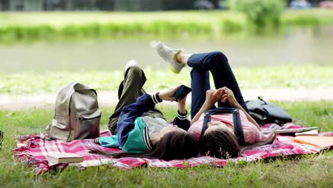 Tracking-Aufnahme-von-zwei-entspannten-College-Mädchen-ruhen-auf-Rasen-in-der-Nähe-von-Teich-nach-dem-Unterricht.-Studentinnen-liegen-auf-karierter-Decke,-surfen-in-sozialen-Netzwerken-auf-dem-Smartphone-und-chatten