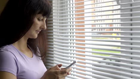 Young--woman-standing-with-smartphone-by-window-in-home.-Close-up,-serious-girl-looking-out-the-window-uses-a-smartphone.Text-message,-online-surfing,-app,-social-network.