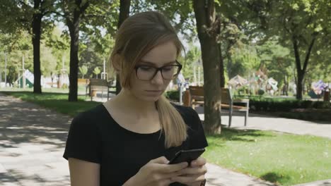 Perplexed-caucasian-woman-in-eyeglasses-walking-checking-smart-phone-content-and-then-stops-looking-at-camera-in-a-park