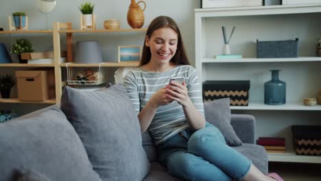 Hübsche-Mädchen-mit-modernen-Smartphone-berühren-Bildschirm-lachen-auf-dem-Sofa-zu-Hause