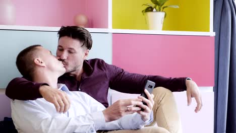 Gay-couple-using-phone-on-arm-chair-together.-Showing-ring-via-video-call-and-kiss.