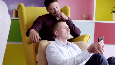 Gay-couple-using-phone-on-arm-chair-together.-Trying-to-selfie.