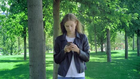 Front-view-of-a-perplexed-woman-walking-checking-smart-phone-content-and-then-stops-looking-at-camera-in-a-park.-A-young-girl-in-street-clothes-looks-at-her-smartphone-in-surprise.-Shocking-message