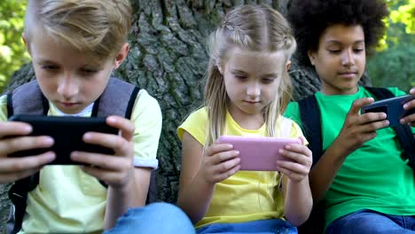 Kinder-spielen-Smartphone-Spiele-sitzen-unter-Baum-im-Park,-Gadget-Sucht