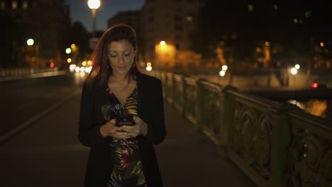 Feliz-atractivo-elegante-mujer-moderna-caucásica-usando-vestido-de-flores,-chaqueta-negra-y-pelo-rojo-caminando-por-la-calle-y-escribiendo-un-mensaje-de-texto-en-su-teléfono-inteligente-por-la-noche.-Paris-4K-UHD.