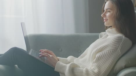 Schöne-junge-Frau-arbeitet-auf-Laptop-Computer,-während-auf-dem-Stuhl-sitzen.-Sinnliche-Mädchen-tragen-Pullover-funktioniert-auf-Notebook;-Studien,-Surfs-Internet,-Nutzt-Social-Media-beim-Entspannen-in-Cozy-Apartment