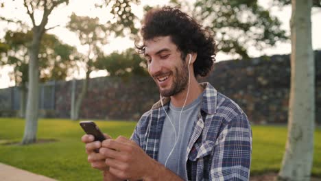 Retrato-de-un-joven-guapo-feliz-usando-auriculares-en-los-oídos-disfrutando-de-mensajes-de-texto-en-el-teléfono-inteligente-en-el-parque---hombre-feliz-usando-su-móvil-en-el-parque