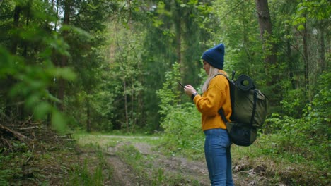 Slow-motion:-Young-woman-hiker-using-her-smart-phone-while-enjoying-her-weekend-in-forest.-Traveling-woman-with-backpack-walking-on-path-the-forest-looking-at-the-telephone-and-map-in-green-wood