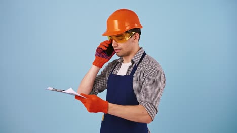 Young-male-in-safety-clothes,-hard-hat,-protective-goggles,-gloves.-Talking-by-cellphone,-looking-at-papers-on-clipboard.-Posing-on-blue-background