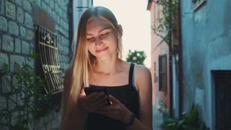 Close-up-of-positive-blonde-woman-reading-something-on-smartphone-while-walking-the-street