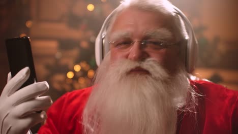 actor-in-image-of-Santa-Claus-is-listening-to-music-by-wireless-headphones-and-smartphone,-closeup-shot-of-funny-face