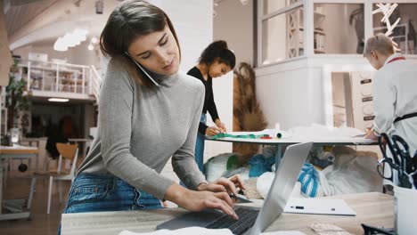 Mujer,-gerente-del-taller-de-costura-aclarando-los-detalles-del-orden-con-el-cliente-por-teléfono-inteligente,-escribiendo-en-el-ordenador-portátil,-sastres-de-carreras-mixtas-trabajando-cerca